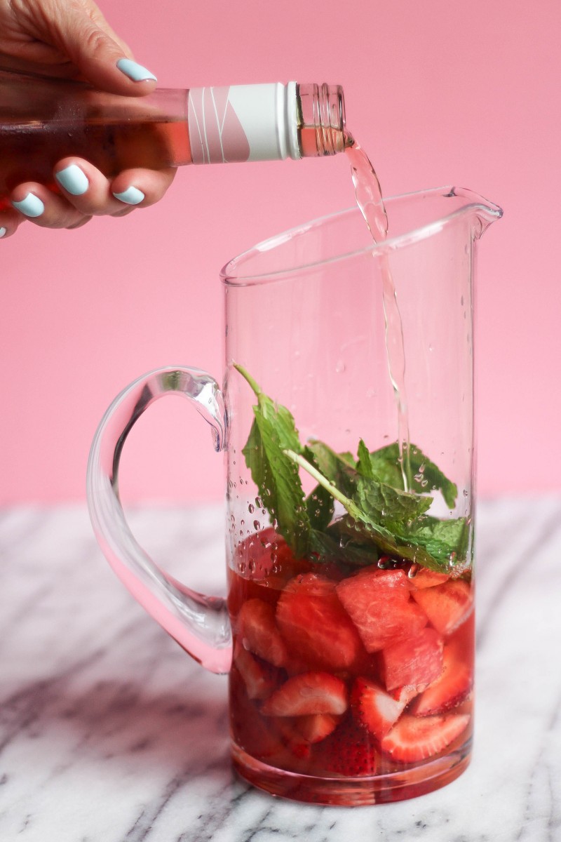 Wine being poured into a tall pitcher filled with watermelon, strawberries and mint.